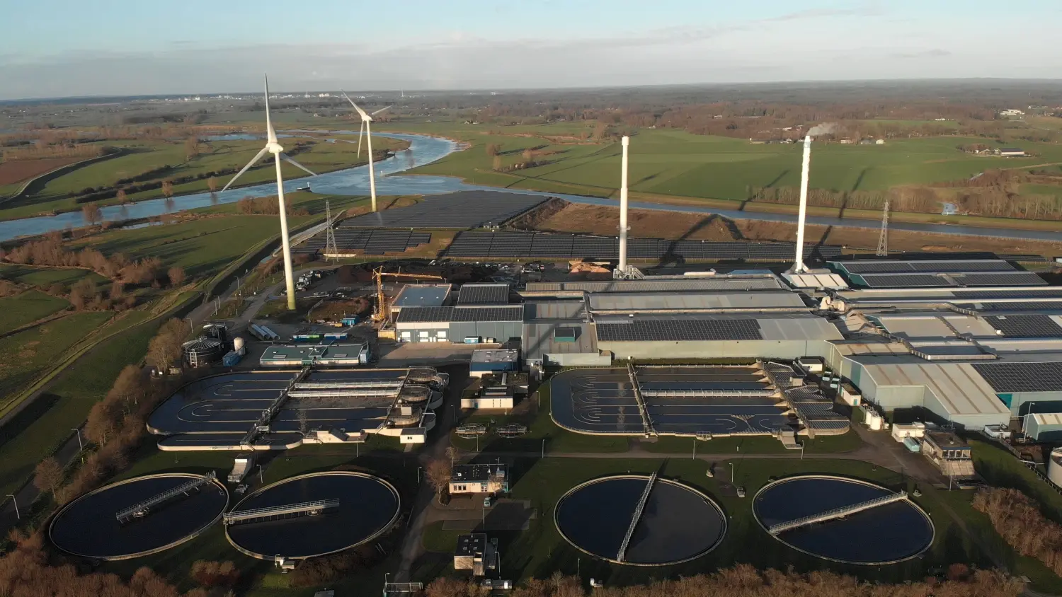Energieberatung - Energieeinkaufsmanagement -Wasseraufbereitungs- und Bioenergieanlage, Windkraftanlagen und Solarpaneele in den Niederlanden sind Teil einer nachhaltigen Industrie in einer flachen Flusslandschaft vor blauem Himmel. Konzept der Kreislaufwirtschaft aus der Vogelperspektive.